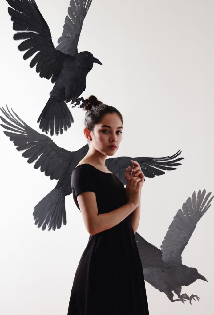 Maria Fernanda in a black dress stands against a white background with three black birds in various stages of flight around her.