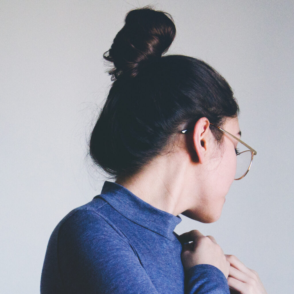 Maria Fernanda with a top-knot hairstyle and glasses is captured in a side profile wearing a blue turtleneck sweater.