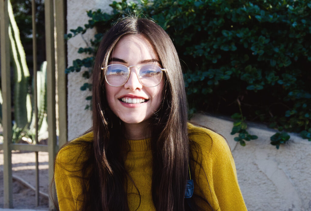 Maria Fernanda with long brown hair and glasses is smiling wearing a yellow sweater against a backdrop of green plants and a beige wall.