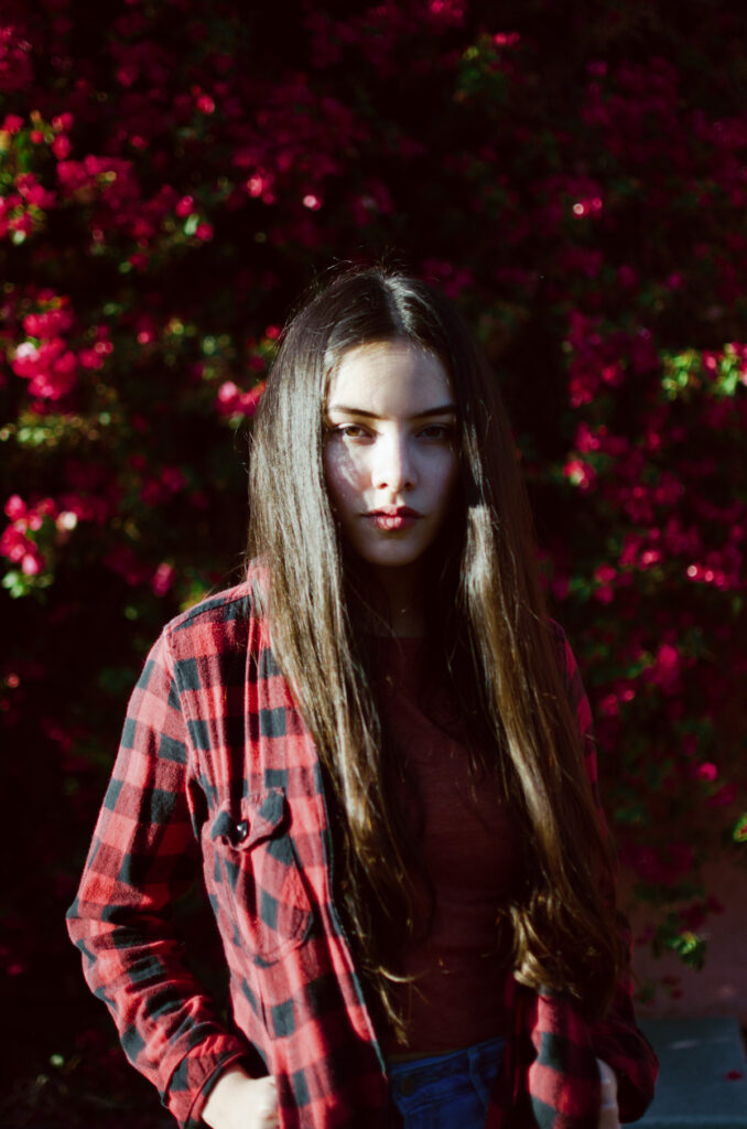 Maria Fernanda with long brown hair stands against a backdrop of vibrant red flowers wearing a red and black checkered shirt.
