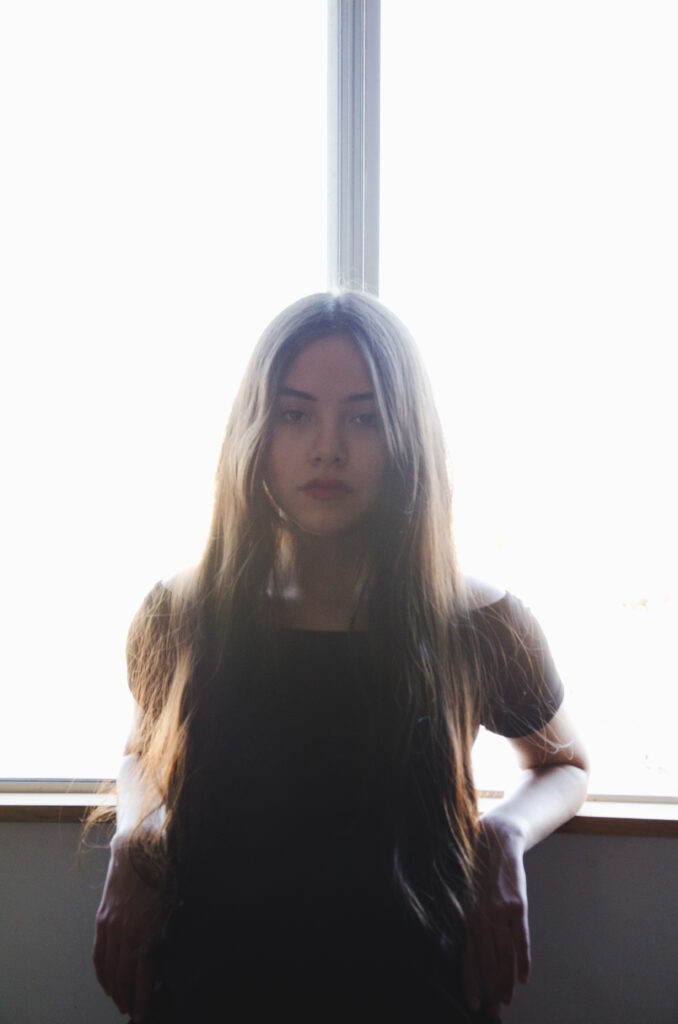 Maria Fernanda with long brown hair stands by a window illuminated by soft sunlight wearing a black top.