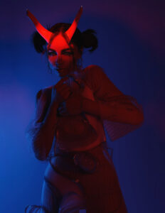 Darian Rojas with red-lit horns and intricate makeup stands against a blue-purple gradient backdrop wearing a translucent outfit with layered wavy designs.