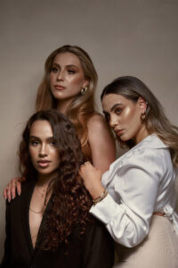 Valeria Clarisa and Abril are three women posing together each showcasing unique beauty and style. They are dressed in elegant attire with Abril in a black blazer Clarissa in a white blouse and Valeria in a beige outfit. Their makeup is sophisticated with a focus on their eyes and lips.