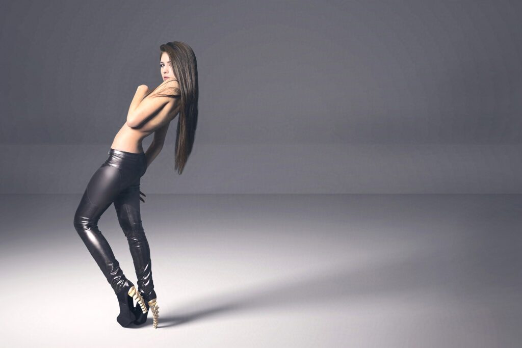 Sarah Cruz with long hair stands in a minimalist setting wearing shiny black leggings and unique heels.