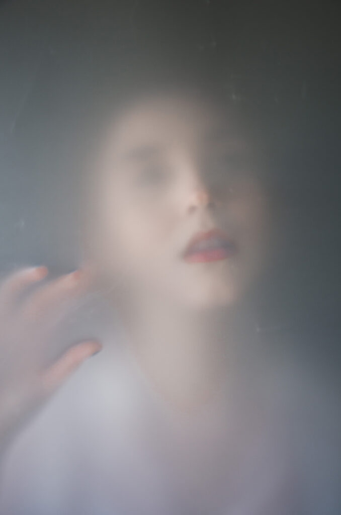 Miranda Valdez's ethereal portrait captures her face obscured by a soft diffused haze with her hand gently touching the glass emphasizing her delicate features and the subtle shimmer of her red lipstick.